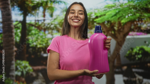 Tableau sur toile Woman holds large pink detergent bottle with both hands in forest setting presenting product; cleaning joy home care