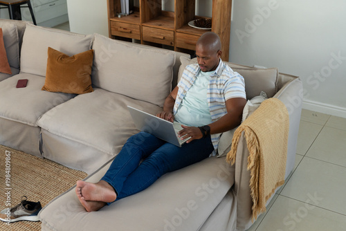 African American man sitting on right section of sofa, using silver laptop at home, copy space