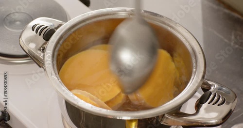 Spoon entering frame is stirring round orange slices in stainless pot on stove, preventing sticking