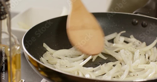 Wooden spatula entering frame is stirring sliced white onions in nonstick pan to soften and glaze