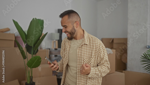 Quadro su tela Man holding smartphone smiling with fist pump in building stacked with moving boxes and houseplant; moving day joy