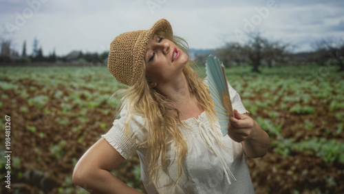 Εκτύπωση καμβά Woman fanning herself with a folding fan in forest field wearing straw hat and white blouse, hand on hip and eyes closed; relief cooling