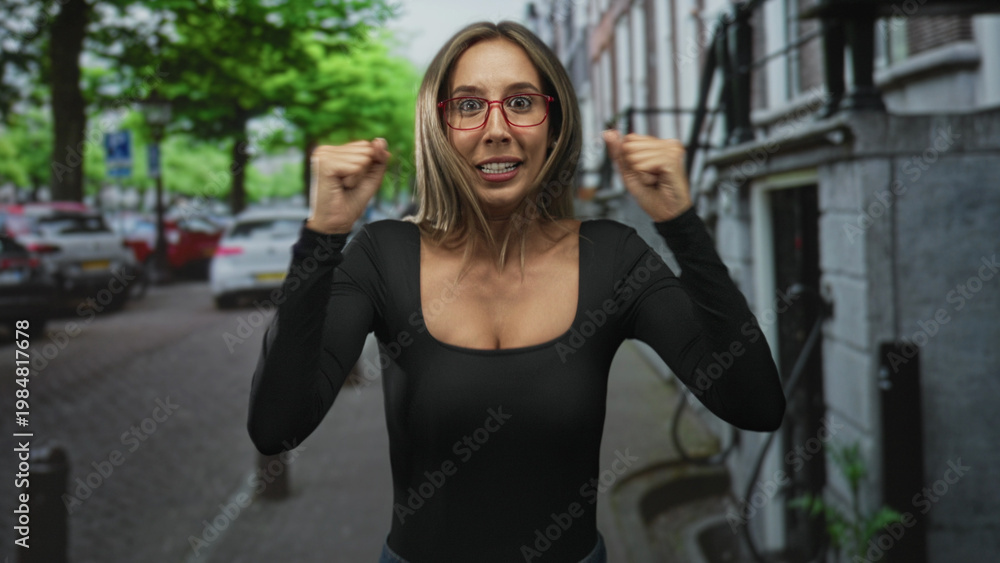 Obraz premium Woman wearing red glasses, smiling with fists raised and visible cleavage on a busy city street; victory celebration.