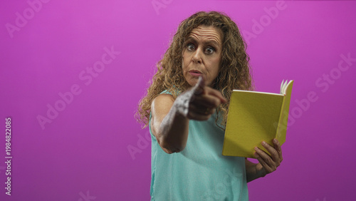 Middle aged hispanic woman pointing finger and holding yellow book in studio with magenta backdrop; reproach warning.