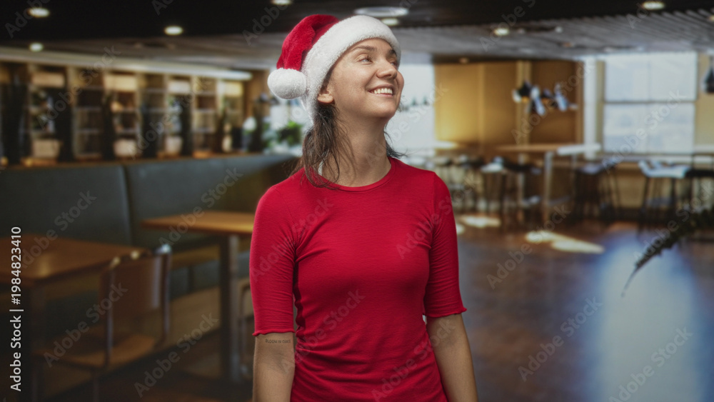 Obraz premium Woman wearing santa hat smiling and looking up inside a building cafe with wooden tables and chairs; cheerful holiday.