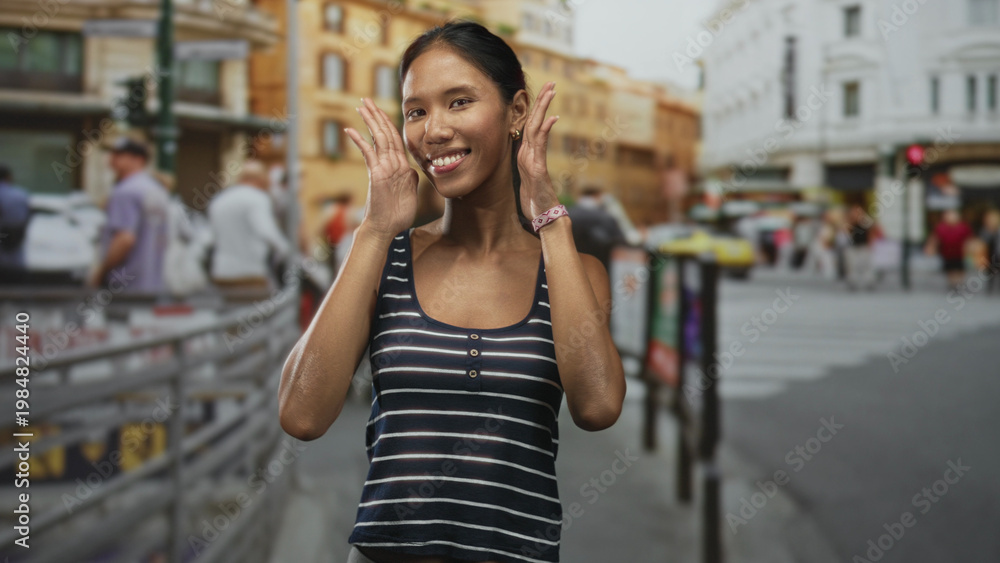 Fototapeta premium Thai woman waving hands and smiling with face visible on a busy city street near buildings and crosswalk, wearing striped tanktop; travel joy.