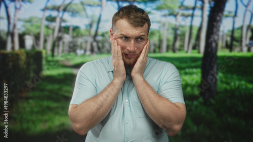 Man pressing hands to cheeks and frowning on a tree lined forest path, standing amid green park trees; concern.