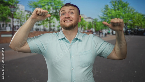 Schilderij op canvas Man in light blue shirt pointing thumbs to chest on a city street, smiling widely with beard and raised arms; pride