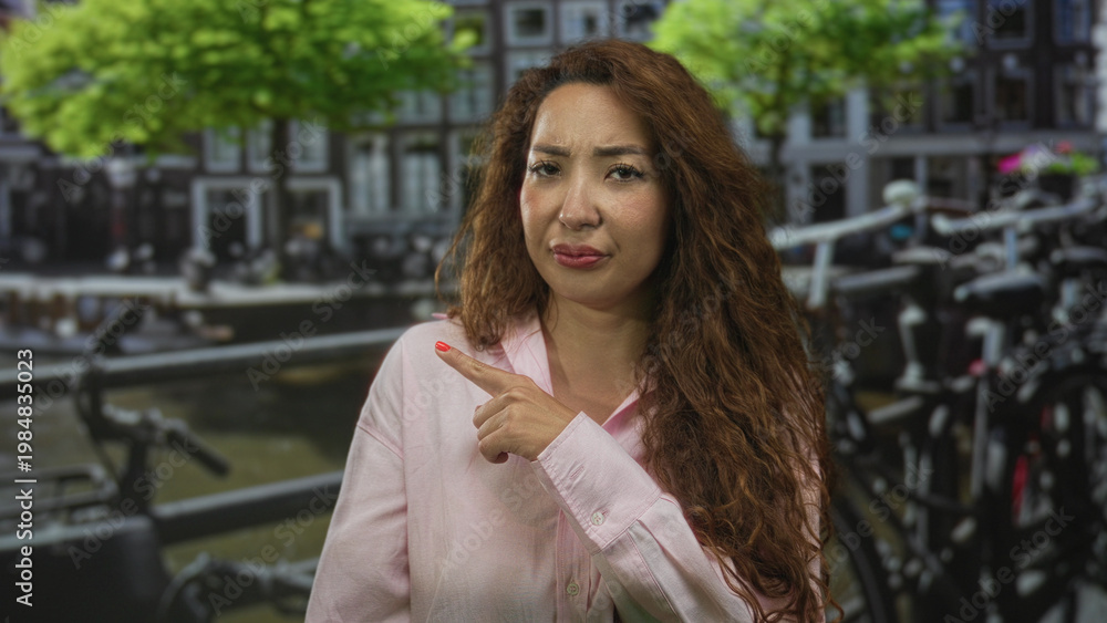 Fototapeta premium Woman pointing finger on street beside a canal in amsterdam with parked bicycles and building facades visible, frowning expression; doubt.