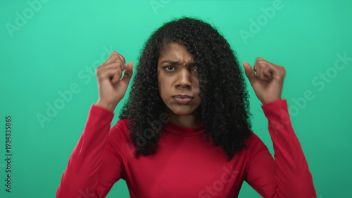 Bild auf Leinwand African american woman with curly hair frowns and raises hands while staring at camera in green studio set; frustration