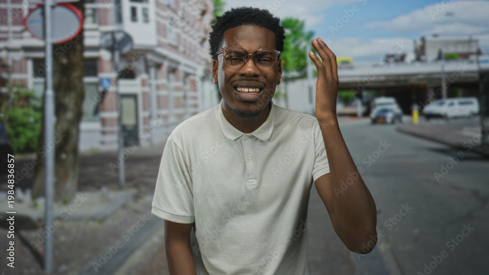 Fototapeta premium Young african american man wearing glasses raises hand to head on busy city street amid cars; confusion.