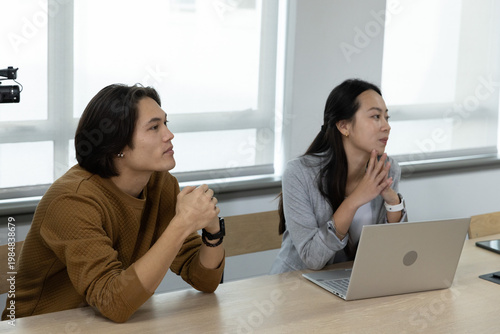 Asian coworkers sitting and listening at wooden meeting table in office with blazer, laptop, camera