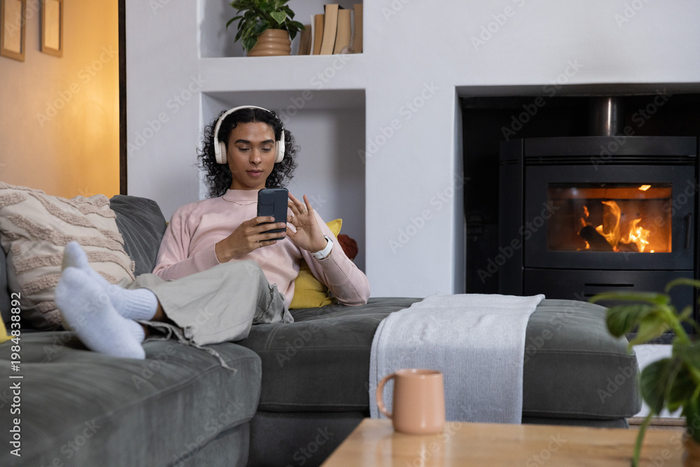 Obraz premium Nonbinary adult reclining on gray sofa at home wearing headset holding phone fireplace glowing