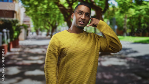 Young black man with glasses making a phone hand gesture by ear wearing a yellow sweater on a tree lined street; friendly connection.