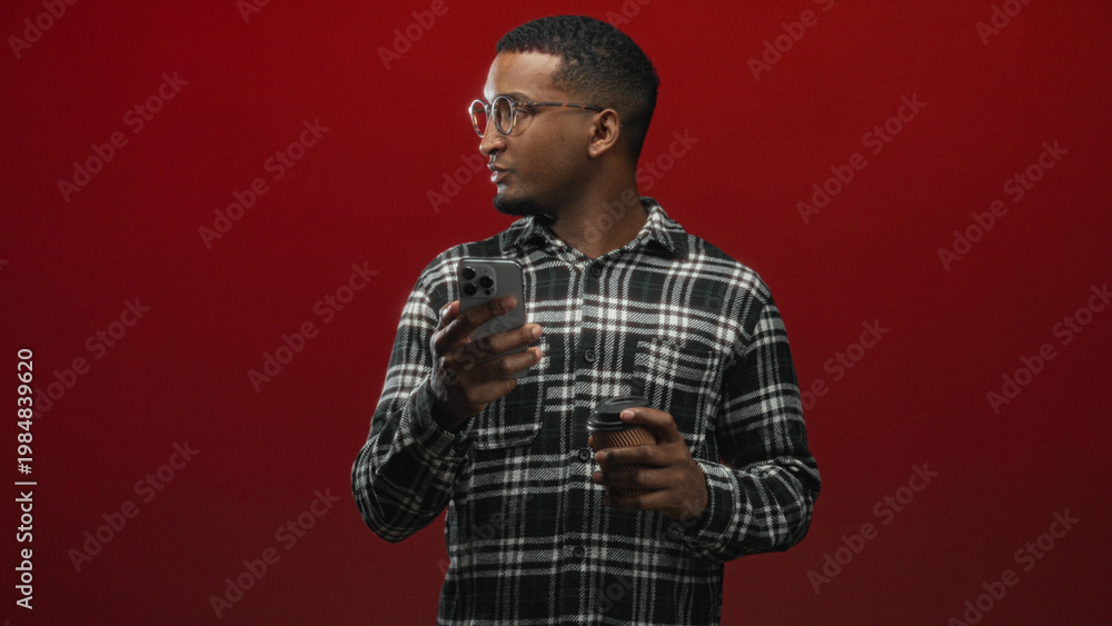 Obraz premium Man with glasses in plaid shirt looking down at smartphone while holding a paper coffee cup against a red studio backdrop; concentration morning routine.