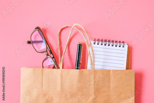 Book note with pen and eyeglasses in shopping bag on color background.