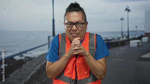 Foto Man in orange reflective vest clasping hands near chest on a street promenade by the seaside with tense face and grimace; unease