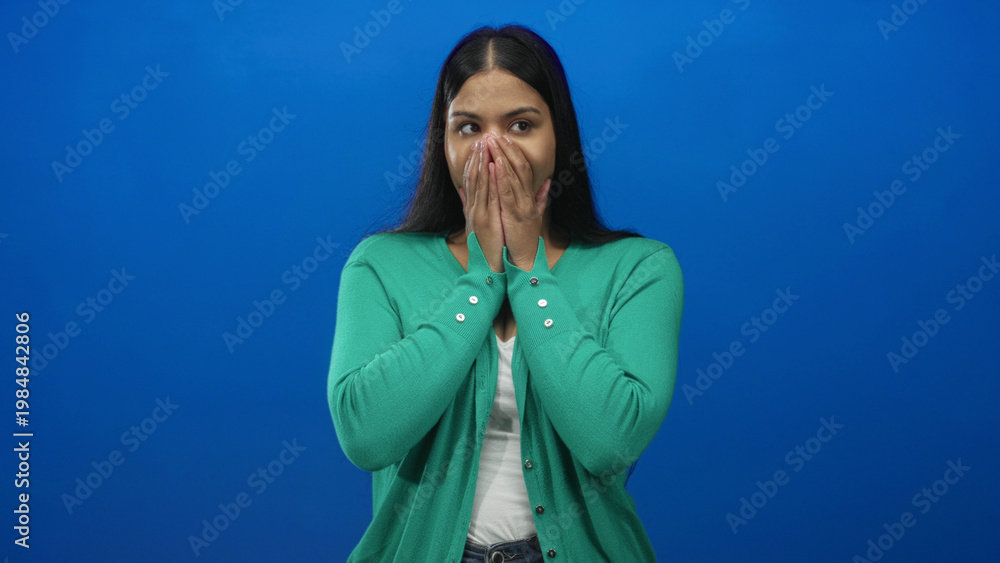 Εκτύπωση καμβά Woman wearing green cardigan covers mouth with both hands fully and closes eyes in blue studio; surprise