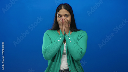 Tablou pe pânză Woman wearing green cardigan covers mouth with both hands fully and closes eyes in blue studio; surprise