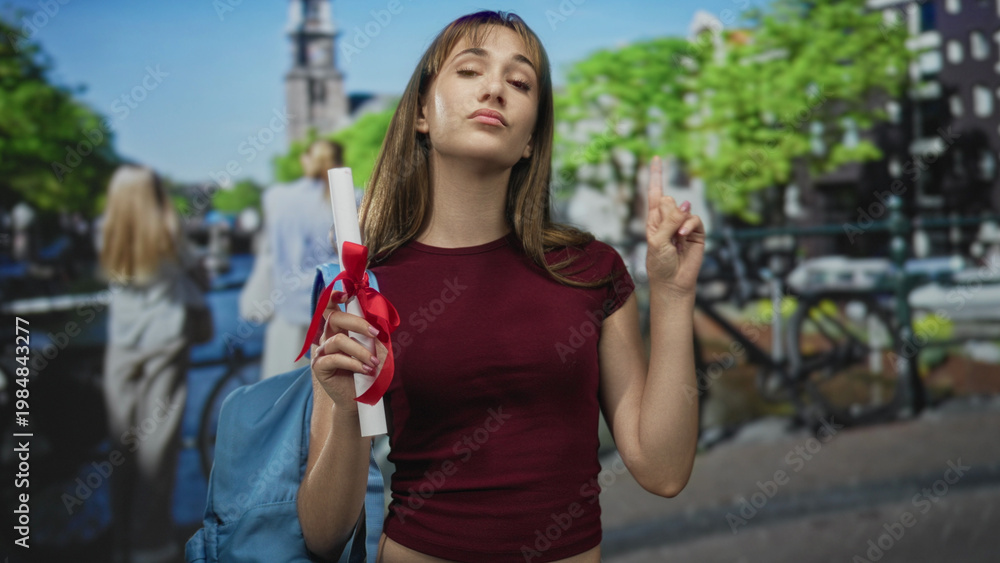 Obraz premium Woman holding diploma scroll tied with red ribbon and blue backpack, pointing finger upward on a street in amsterdam; pride achievement success.
