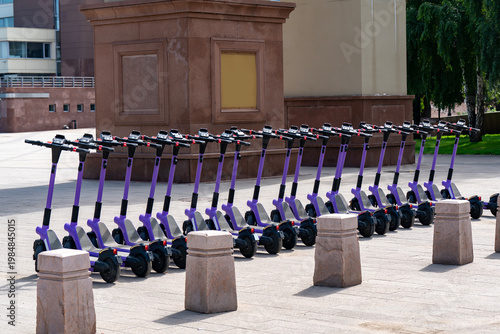 Row Of Purple Electric Scooters Parked In City Square Ready For Rental And Urban Micro Mobility