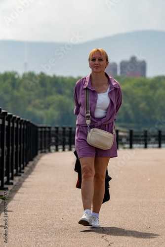 Woman Walking Alone On Riverside Promenade In Casual Purple Outfit With Crossbody Bag