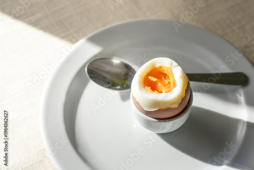 Boiled egg with soft yolk in a stand and a spoon on a white plate on a sunny day, protein snack for breakfast, lunch or dinner, copy space, selected focus