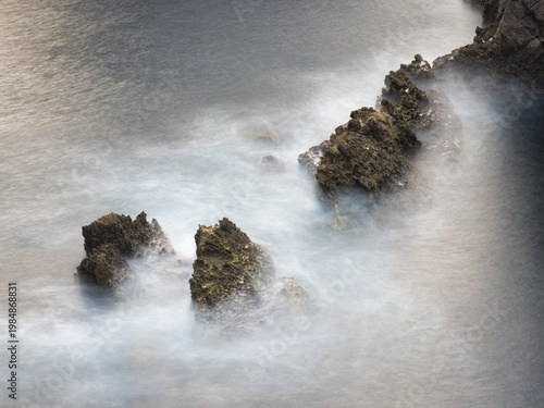 Foto Abstract Jagged Rocks in Mystical Water Motion - High-angle square-format long-exposure of dark, jagged coastal rocks seemingly dissolving in a misty swirl of blurred white and grey ocean water
