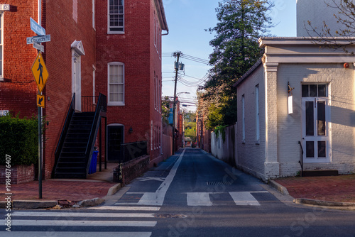 Billede på lærred Scenic narrow alleyway in historic Frederick, Maryland, with classic red brick buildings, a crosswalk, and a yellow pedestrian sign at sunrise