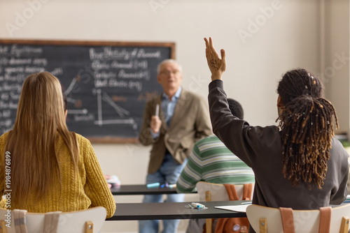 Quadro em tela Black teenage boy raising hand during classroom lesson while senior male teacher explaining economics to students