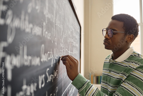 Fényképezés Young African American man writing equations on chalkboard in classroom, studying mathematics and problem solving