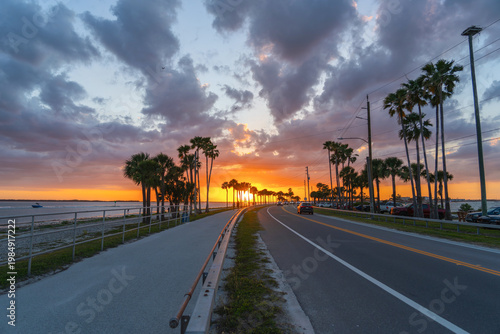 Sunset. Coastal road. Dunedin Causeway Florida. Scenic tropical seaside highway with palm trees along shoreline during colorful sunset sky evening travel landscape. Cars, ocean horizon dramatic clouds