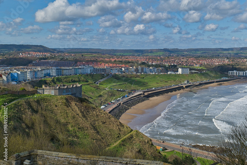 An elevated view captures a coastal town with a sandy beach, breaking waves, and buildings along the cliffs. A fort-like structure sits on a green hill  in Scarborough Castle - Yorkshire - Great Brita