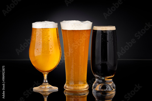 Three glasses of beer including lager, wheat beer and dark stout on black background with foam and condensation. Cold refreshing alcoholic beverages in commercial product photography style.