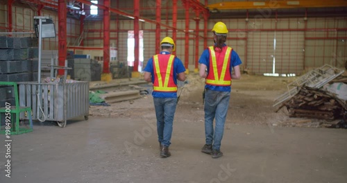 Two industrial workers in safety gear walk through a large factory warehouse, collaborating on a site inspection for operations and quality control assessment.