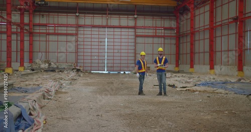 Two engineers in hard hats inspect a demolition site inside a large, abandoned warehouse, planning the next phase of an industrial redevelopment project.