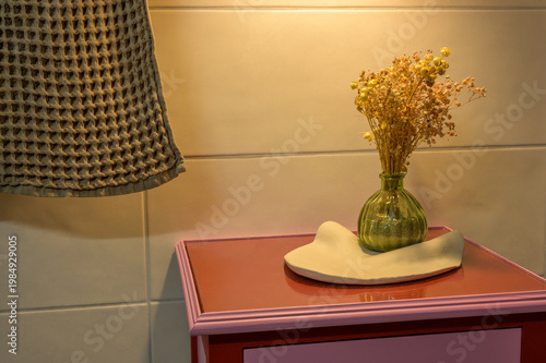 Dried flowers in green vase on white pottery stand on pink nightstand with checkered lampshade in soft indoor lighting. Cozy indoor furniture.