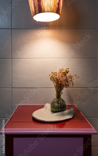 Dried flowers in green vase on white pottery stand on pink nightstand with checkered lampshade in soft indoor lighting. Cozy indoor furniture.