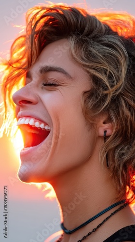 A young man with curly hair laughs heartily with his eyes closed, bathed in the warm, orange light of a sunset