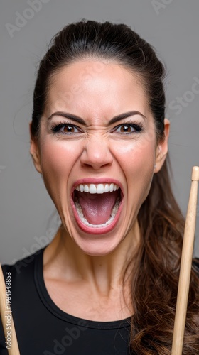 Young female drummer intensely screams with a wide open mouth and furrowed brow, holding drumsticks. Her dark hair is pulled back, wearing a black top against a simple grey studio background