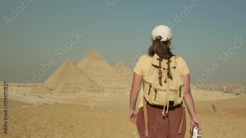 Caucasian woman tourist with backpack walking towards pyramids in Giza on sandy hill clear sunny day, visitor exploring sights of ancient Egypt in Cairo