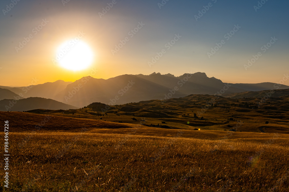 Fototapeta premium Golden hour sun setting over mountain landscape and grassy field