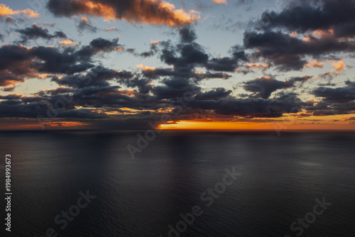 Dramatic sunset over calm ocean with dark clouds and golden light on horizon. Sun rays breaking through clouds create atmospheric seascape scene