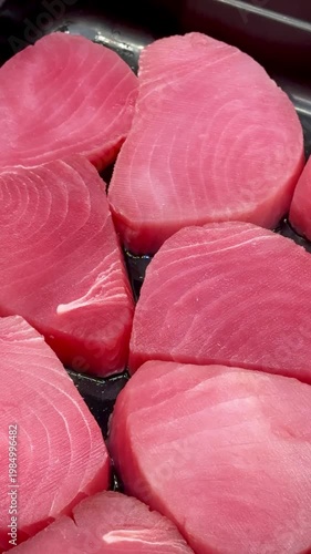 Pink tuna steaks on black tray, supermarket shelf closeup, chilled display of raw fresh fillets arranged in neat rows, subtle.