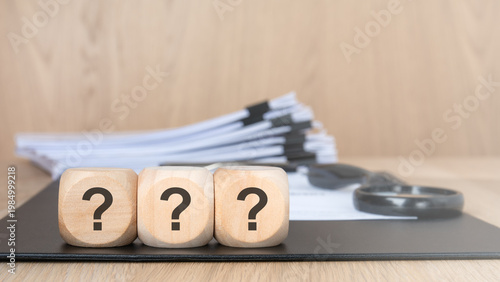 Three question marks on wooden blocks. Blurred background with a document stack and magnifying glass. Corporate audit and investigation concept.