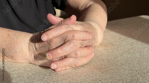 Sunlit elderly hands clasped on table, close-up showing wrinkled skin, prominent veins, gentle manicure, warm late afternoon light.