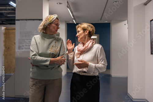Women standing in gallery interior examining framed artwork and pointing during exhibition visit in quiet discussion.
