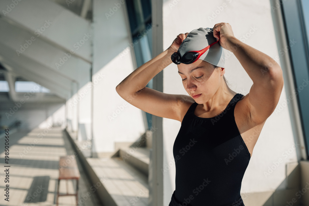 Naklejka premium Young woman athlete preparing for her swimming workout, putting on a swimming cap and goggles in an indoor pool facility