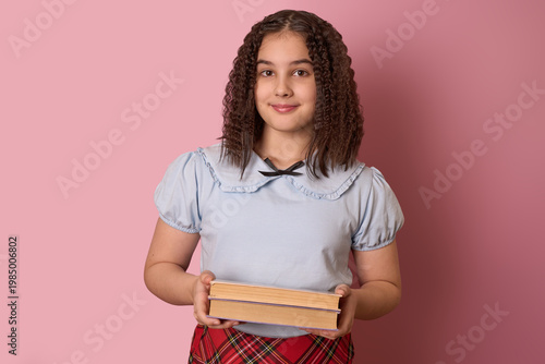 Young Girl with Curly Hair Holding Books Against a Soft Pink Background Smiling Brightly