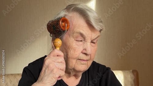 Close-up senior woman massaging scalp with magnetic needle roller, focused expression, wooden handle contact, soft sunlight.